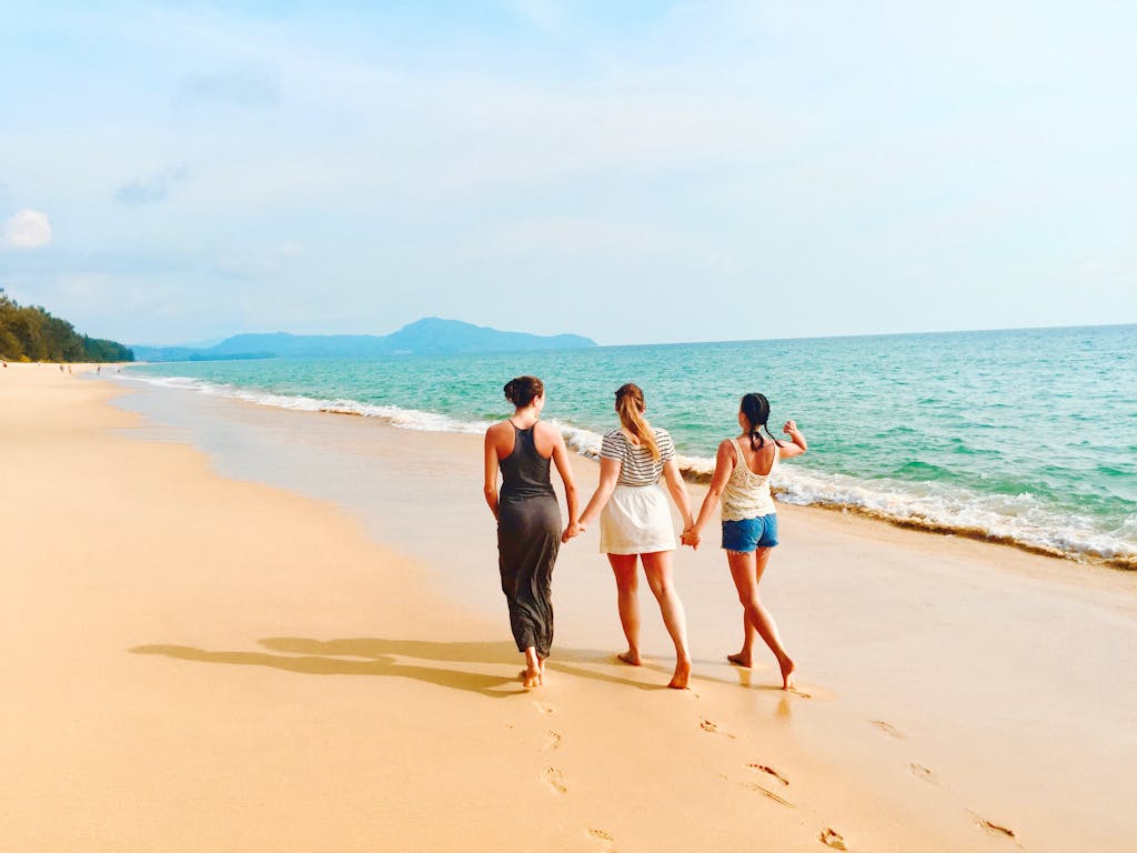 Three women walking hand in hand on a sandy beach in Thailand with waves and horizon.